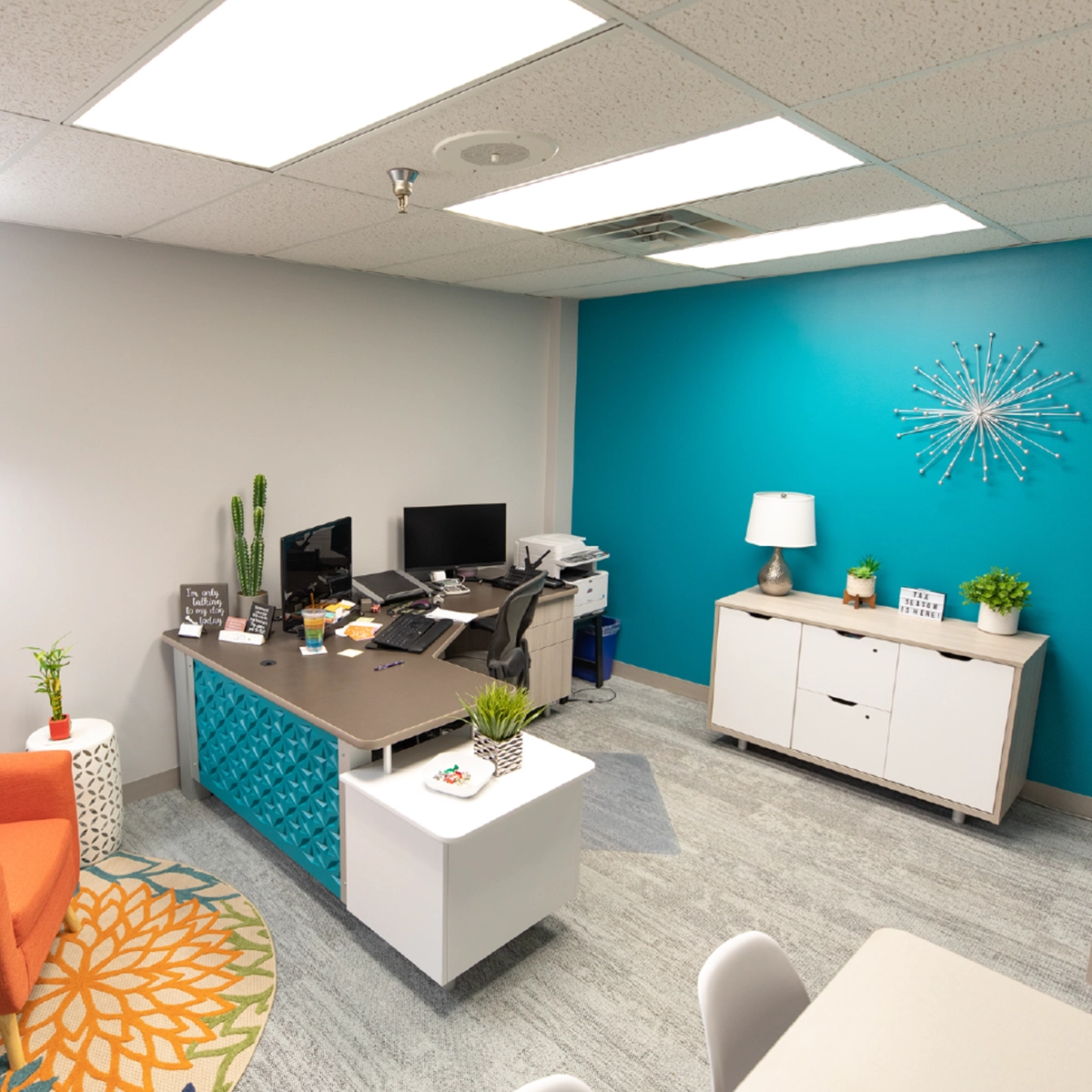 A neatly organized modern office space for rent with an L-shaped desk, teal accent wall, decorative starburst art, and white furniture accents under bright fluorescent lighting.
