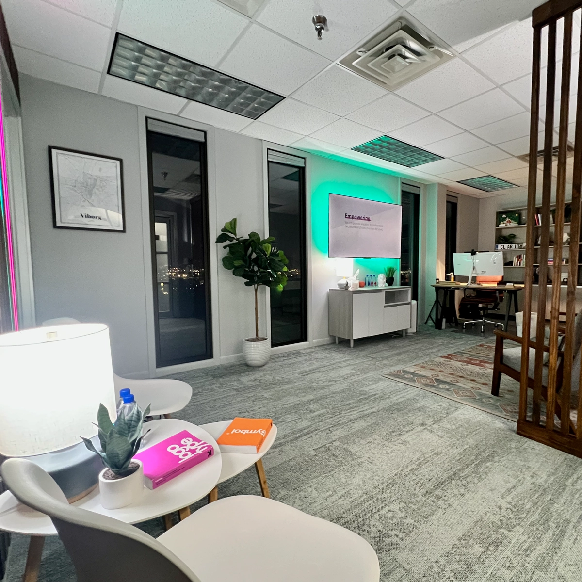 Modern office space for rent with seating area, LED-lit "Empowering" sign on monitor, workstation with dual monitors, and decorative accents like plants and colorful notebooks.